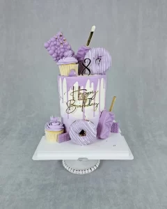A tall white and purple birthday cake decorated with donuts, cupcakes, ice cream, and chocolate